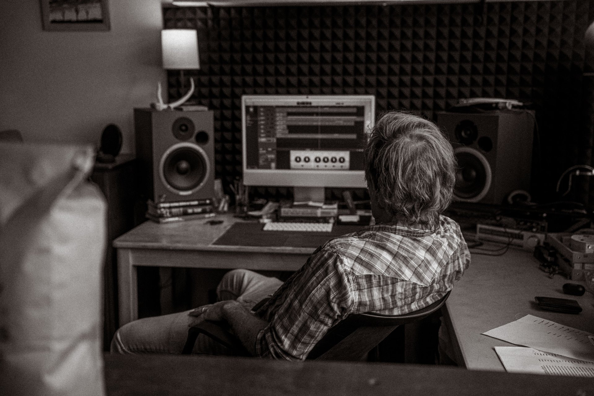 A man seen from behind at a desk, working on an audio track on a computer between two studio speakers in a room with foam panels on the wall.